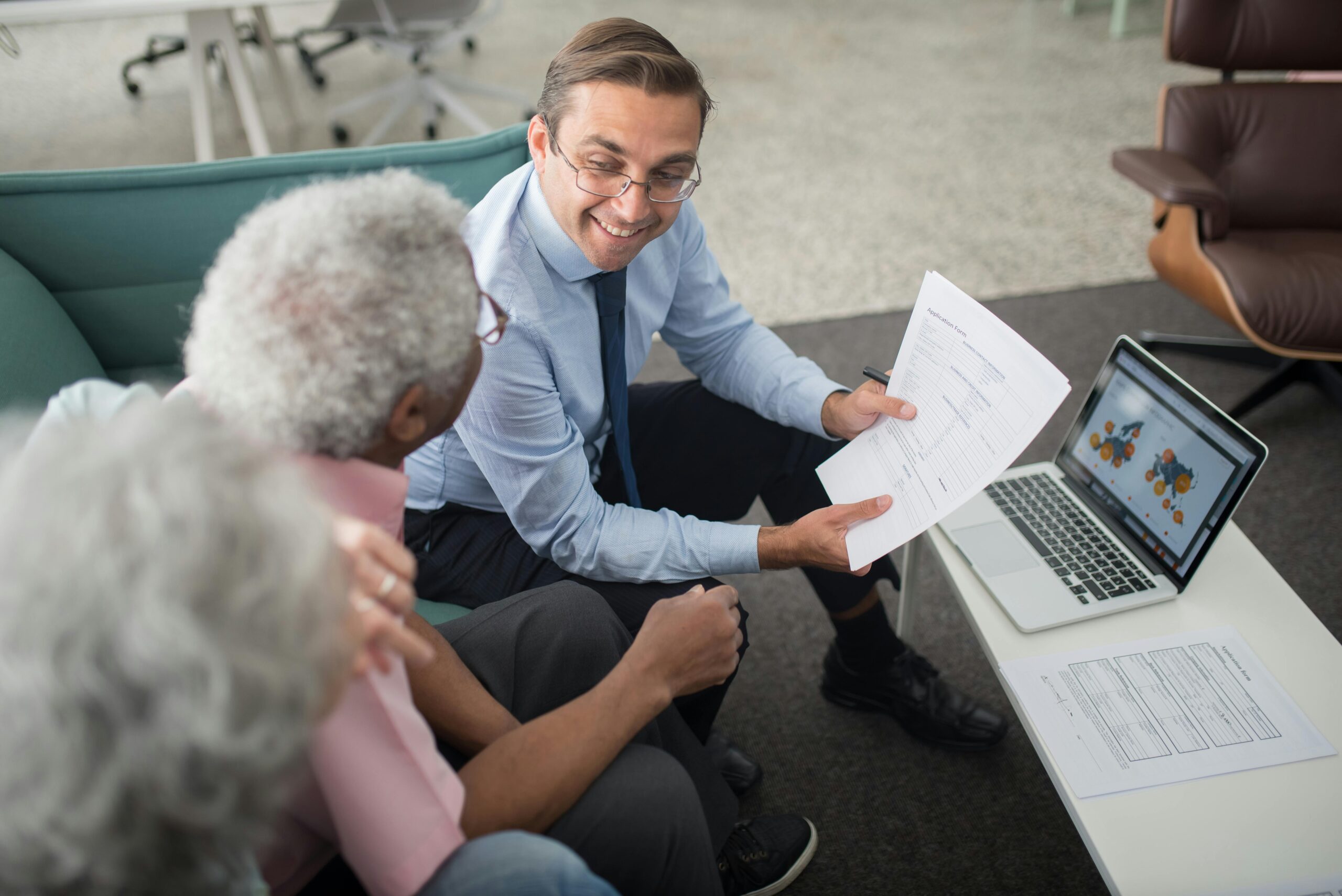 Man discussing funeral insurance options with an elderly couple, holding documents, laptop displaying relevant information, in a comfortable office setting.