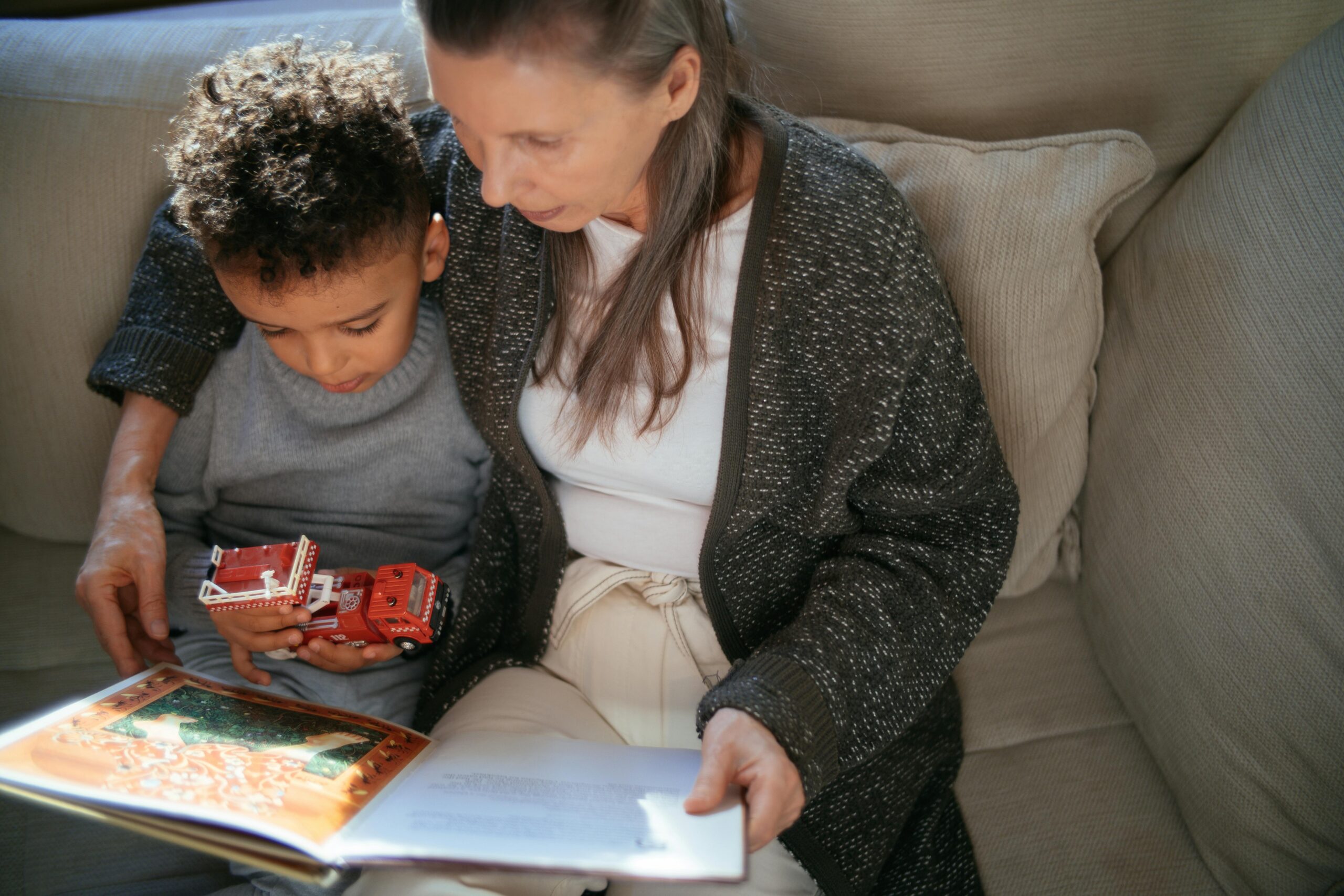 Elderly woman reading a book with a young boy holding toy fire trucks, illustrating family bonding and nurturing relationships.