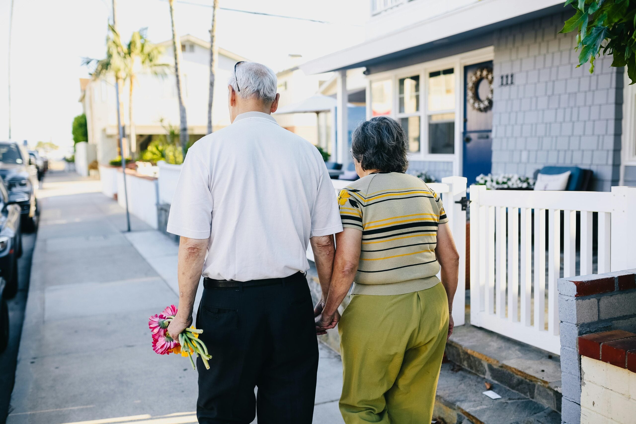 Elderly couple walking hand in hand down a sunny street, man holding a bouquet of flowers, symbolizing love and companionship, relevant to planning for end-of-life care and emotional support in funeral insurance discussions.