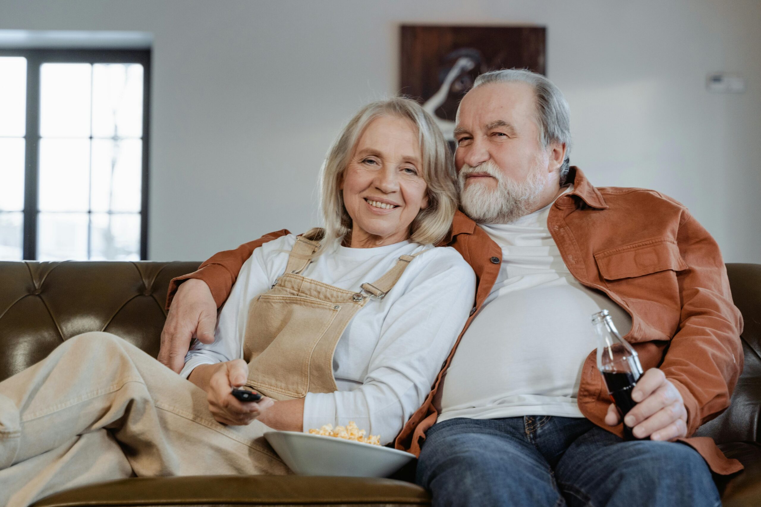 Older couple sitting on a couch, smiling and enjoying time together, with a remote control and a bowl of popcorn, reflecting the theme of family and comfort in planning for end-of-life arrangements.